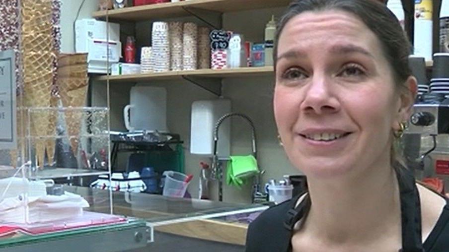 Isabel Morris at the counter of the gelato stall. She has dark tied back hair and is wearing earrings. To her right there is a display of different types of ice cream cones, a sink and various utensils.