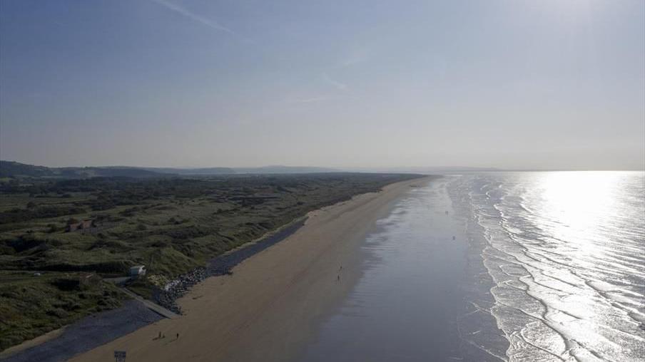 Drone footage of Pendine Sands, showing the 7 mile narrow flat, expanse, from the western most Tenby end towards Carmarthen. People on the beach appear miniscule. On the left is a landscape of shrubbery, whilst on the right the sun is shining on the sea.