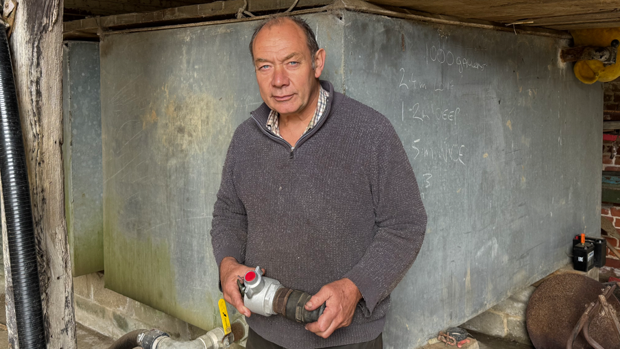 Glenn Buckingham looking directly into the camera. He is wearing a shirt under a piece of knitwear. He is holding a universal coupling and standing in front of two large water tanks.