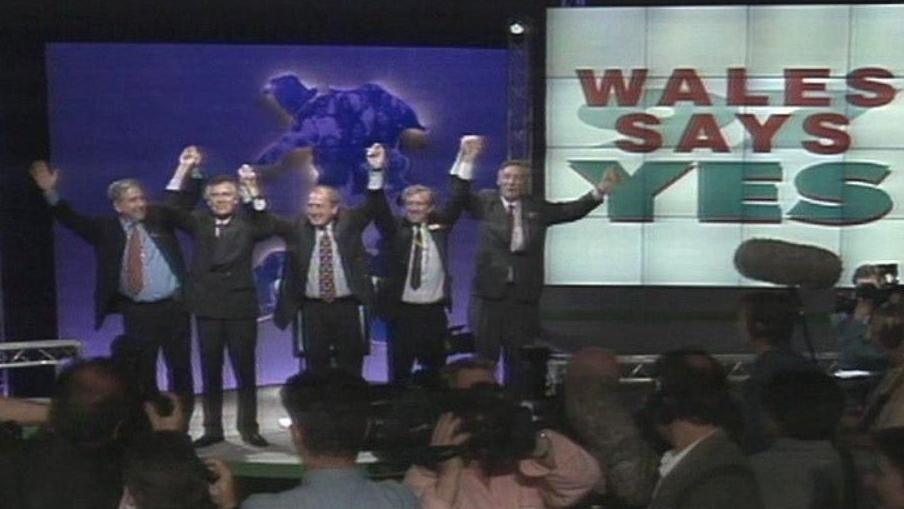 Llun o bedwar o aelodau mwyaf blaenllaw'r ymgyrch Ie yn sefyll ar lwyfan yn dathlu canlyniad 1997. Mae eu breichiau yn yr awyr ac maent yn dal dwylo. Y tu ôl iddynt mae sgrîn gyda'r geiriau 'Wales Says Yes'.