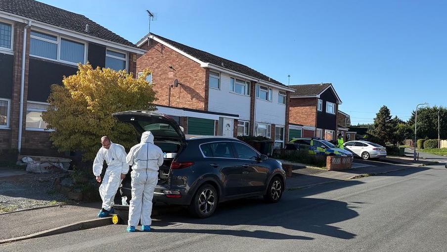 Police in white overalls are outside a house in Oak Apple Close. The road has houses on both sides and cars parked in the street and in driveways. An officer can be seen in a hi-vis jacket further away.