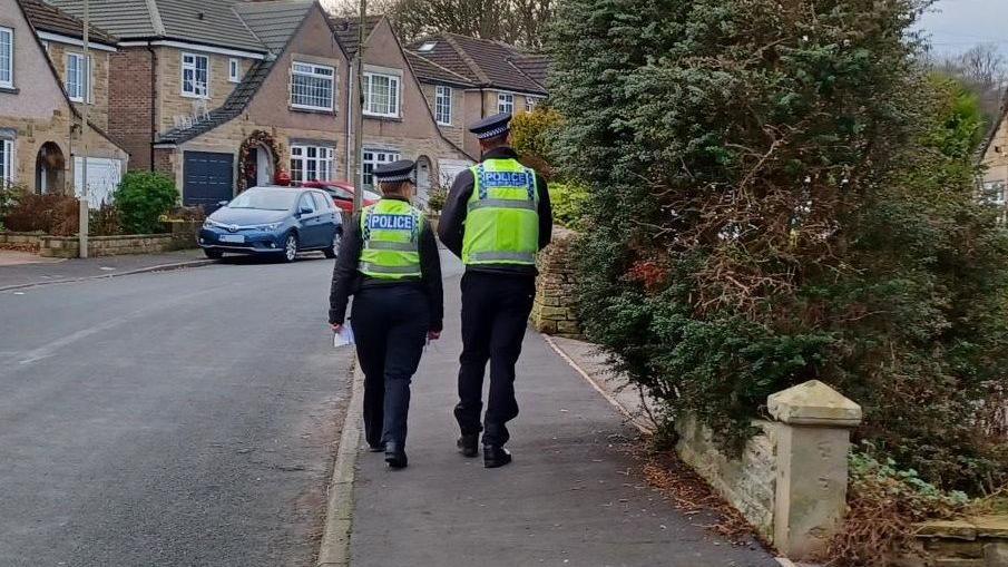 Two officers in police uniform walk along a pavement. Hedges can be seen on the right, while a row of houses can be seen on the left. A blue car is parked in front of one of the houses.