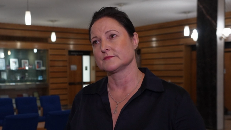 A woman in a black, open-necked blouse with tied back dark hair stands in a wood-panelled room full of blue chairs.