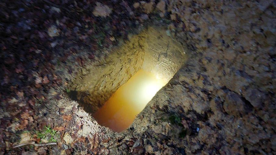 A rectangular hole dug in the ground, with a pool of water at the bottom. It's dark, with the hole appearing to be lit by torchlight.