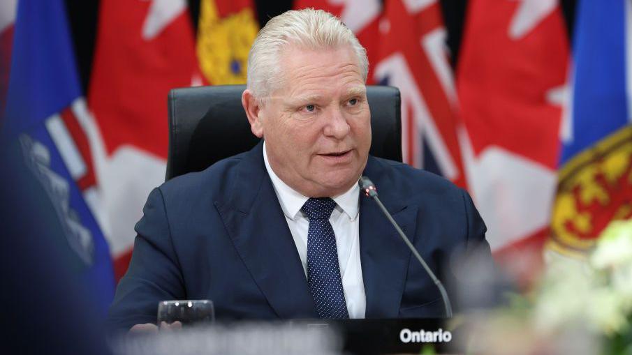 Doug Ford speaks at a meeting in Canada. He is sitting by a desk in front of a row of national flags, including those belonging to the UK and Canada.