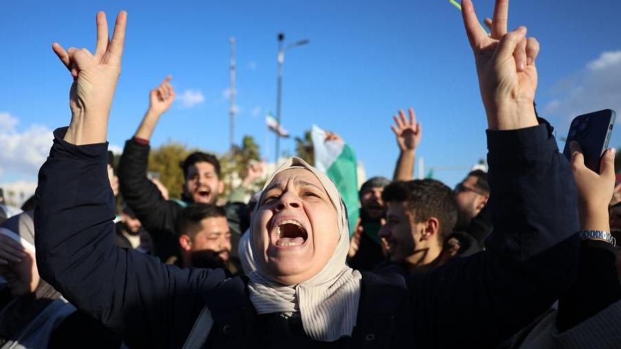 A woman raises both arms and makes the peace sign with her hands in Umayyad Square in Damascus. The sky behind her is a clear blue, and men behind her can be seen celebrating. She looks like she's singing or shouting, with her mouth open and a happy expression.