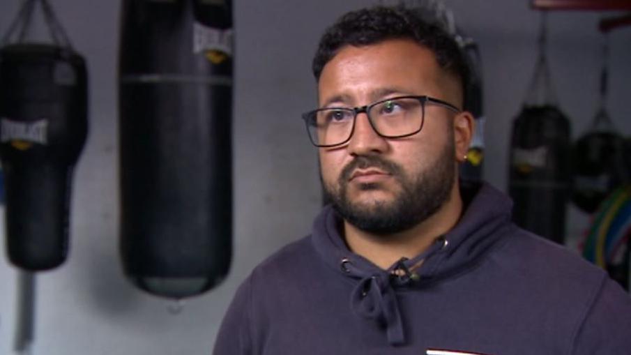 Courtney Young wearing black square glasses. He is wearing a blue hoodie and has black hair with a black beard. Behind him is a boxing gym with long boxing bags.