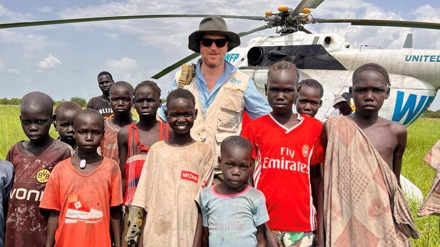 Shaun stands in front of a helicopter in a field, alongside a group of young children in South Sudan. 
