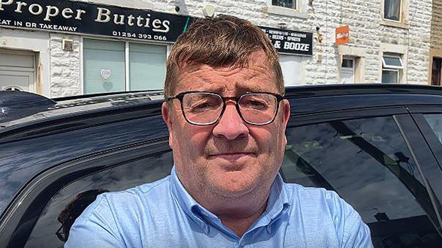 David Fielding with brown hair standing in front of a black vehicle and a sandwich shop wearing black glasses and a blue shirt.