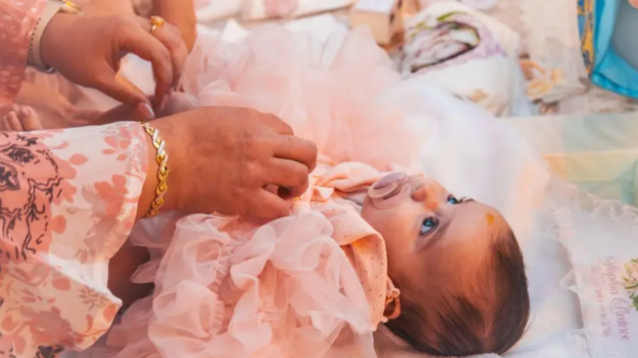 Baby Sophia lies down in a bed. She is wearing pink baby clothes. She sucks a dummy and looks up to the ceiling. A pair of female hands corrects her clothes. The woman wears a gold bracelet and rings.