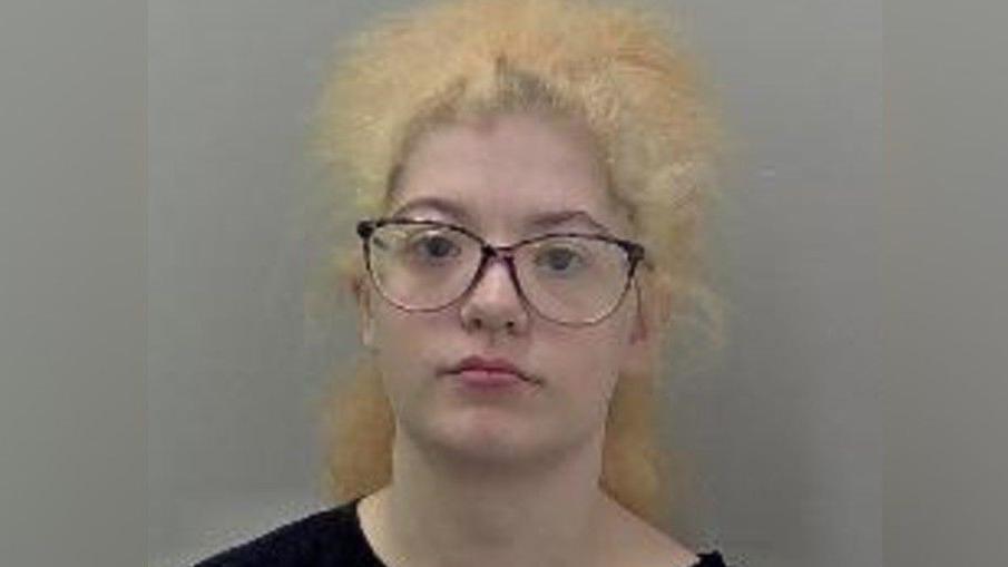 A mugshot of a woman with curly yellow-blonde hair and dark framed glasses. She is wearing a black t-shirt and staring at the camera with a serious expression.