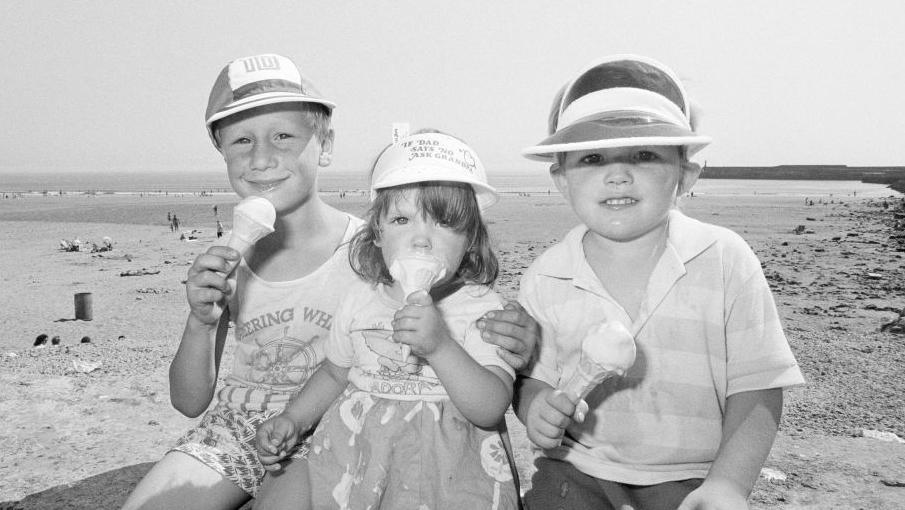 A black and white photo of three children sat on a beachside wall, all wearing hats and holding ice creams. Behind them is the sandy beach and sea.