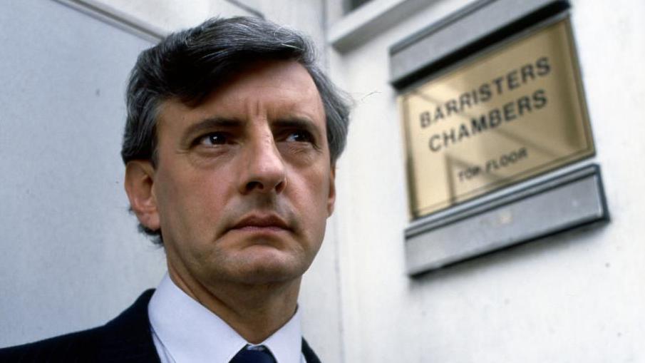 A man in a dark suit, white shirt and dark tie stands outside an office.