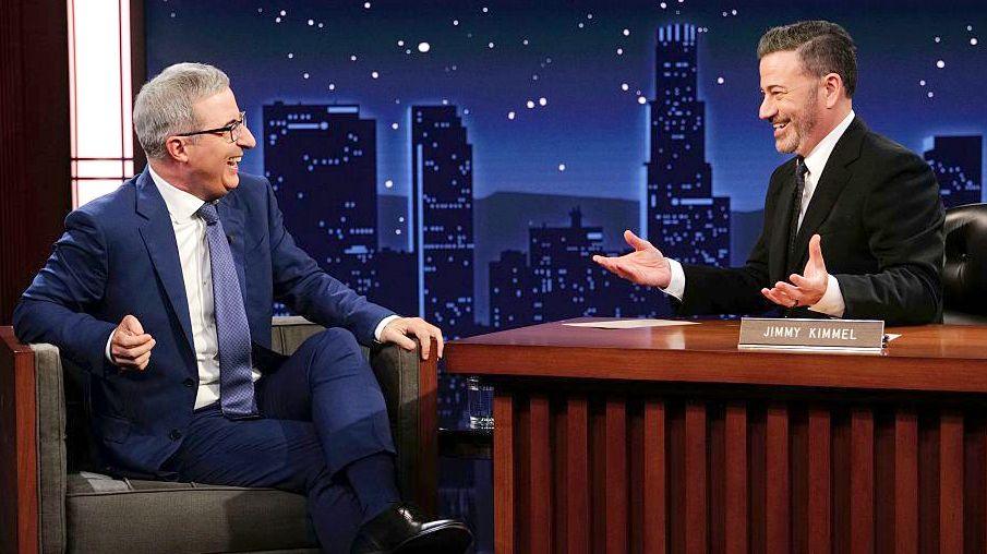 John Oliver sits with Jimmy Kimmel on set during a talk show, they are both smiling and sitting against a night sky cityscape backdrop