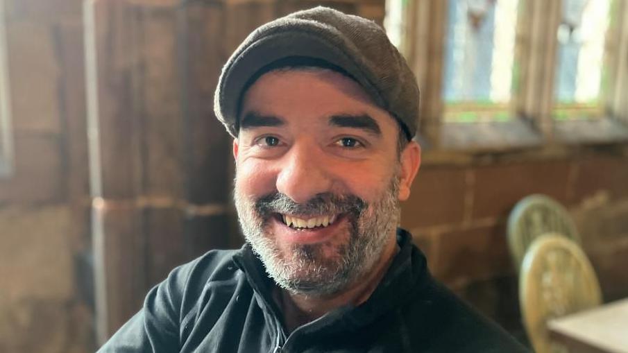 A headshot of a man, who is sitting in a cathedral smiling at the camera. He is wearing a grey flat cap and has a grey beard.