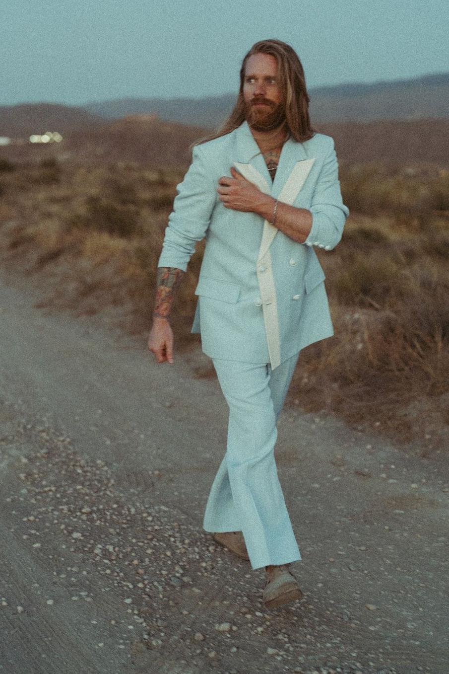 Sam Ryder walks along a gravel path in the middle of the countryside, while wearing a powder blue suit and moccasins