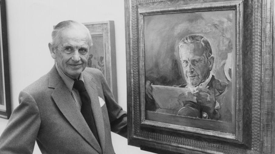 A black and white photo from 1977 of an older Graham Sutherland - a man with short grey hair wearing a suit, striped shirt and tie - stood next to one of his self-portraits in a gallery