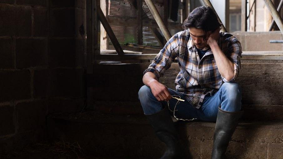 Stock photo of a young farmer sitting with his head in his hands in a barn