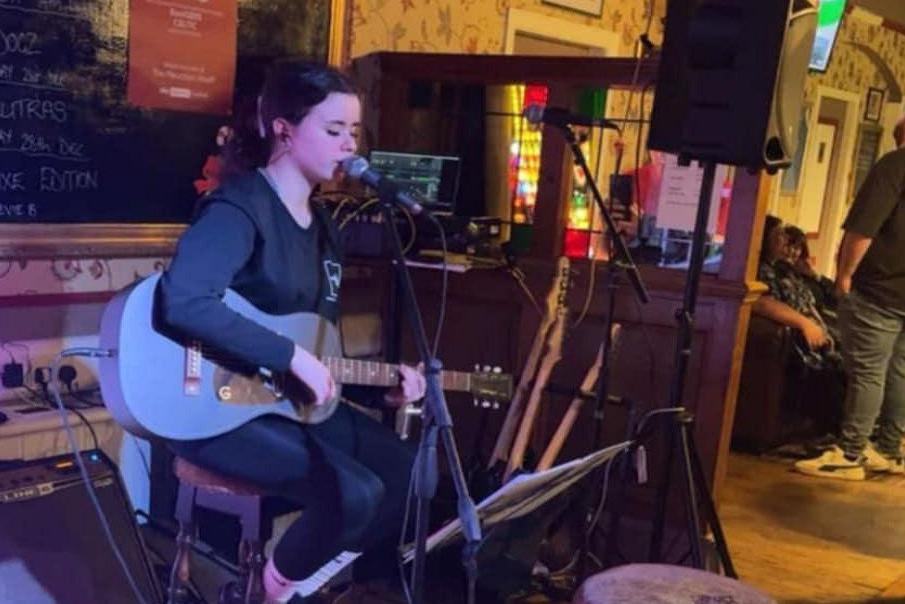 Stevie, a teenage girl with tied-back , dark hair, is sitting on a bar stool playing a blue acoustic/electric guitar and singing into a mic.