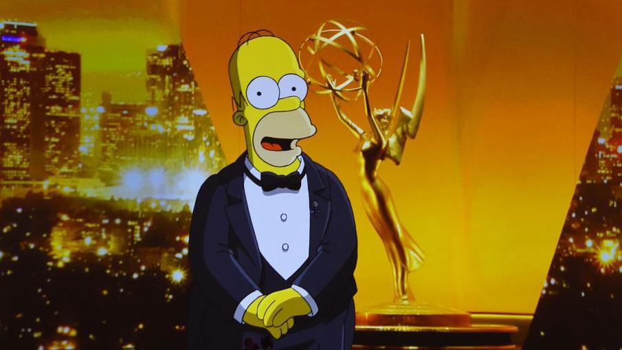 Cartoon character Homer Simpson, who is bald with has yellow skin and stubble, standing with his hands clasped together against a golden background at the Emmy Awards ceremony. He is wearing a black dinner suit, white shirt and a black bow tie. His mouth is open and he looks slightly surprised.