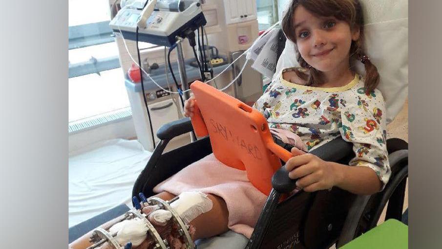 A young girl lying in a hospital bed wearing pyjamas and looking at an ipad with a metal frame on her left leg.