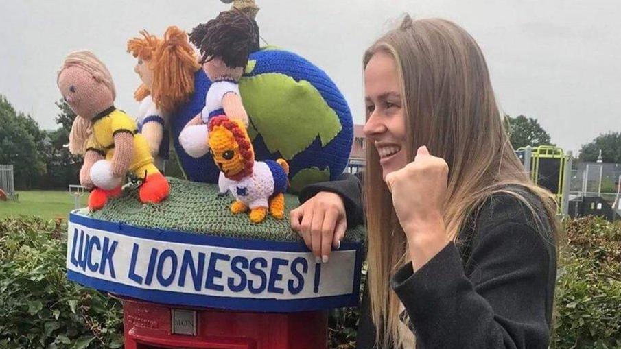 Hannah Hampton next to a postbox topper where part of the wording is displayed and says 'luck Lionesses". On the top are several crafted figures, including a goalkeeper wearing yellow and a lion. Hampton has long blond hair and is making a cheering sign with her hand. 