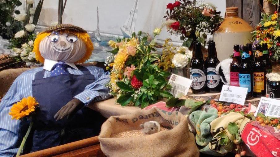 A display includes flowers, bottles of beer, a toy mouse in a sack of grain and a knitted male figure wearing an apron and a cap