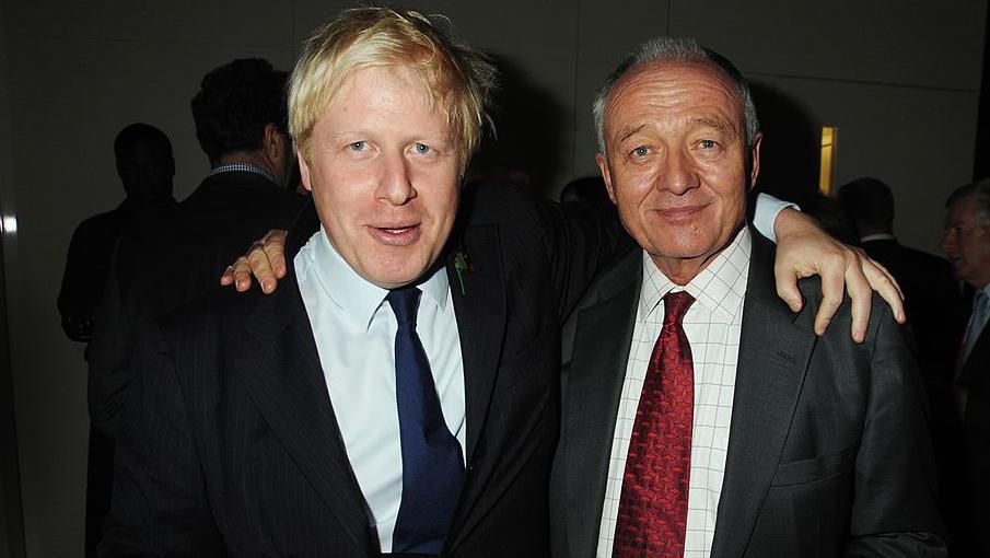 Boris Johnson and Ken Livingstone at a social event with their arms on eachother's shoulders. Both are wearing dark suits, Johnson is wearing a dark blue tie and Livingstone is wearing a red tie. 