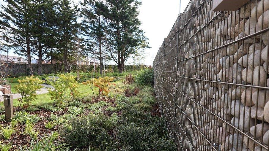 The garden at the new Maggie's centre in Denbighshire. It shows a stone walled garden, young shrubs and trees, a lawned area and a path.