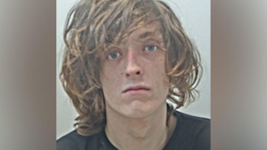 Shane Taaffe Rhodes with long brown unkempt hair, green eyes and wearing a black long-sleeved t-shirt in an image taken by police 