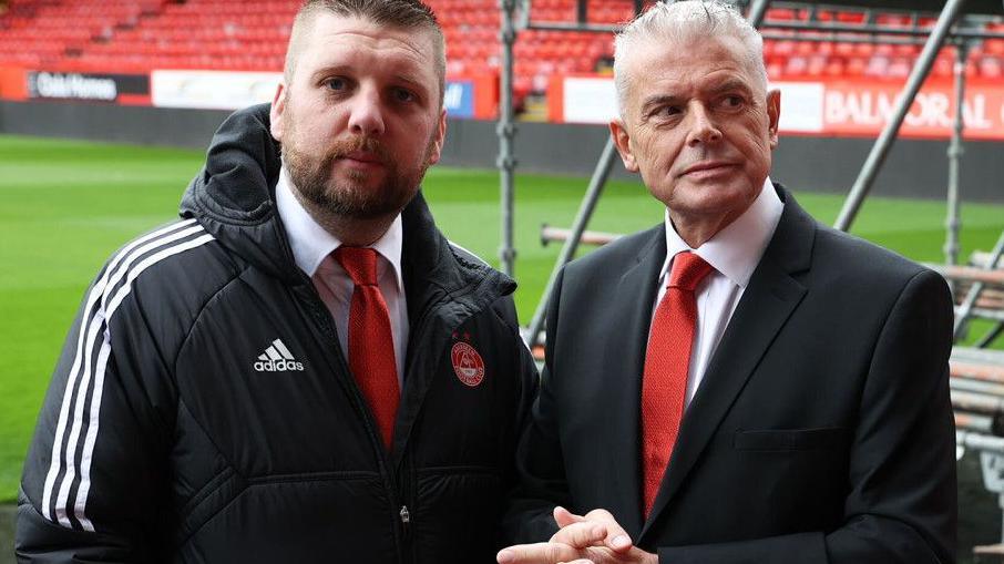 Aberdeen's Alan Burrows and Dave Cormack at Pittodrie. Cormack (left) is wearing a black suit and red tie. Burrows is in a red tie and branded Aberdeen FC jacket. 