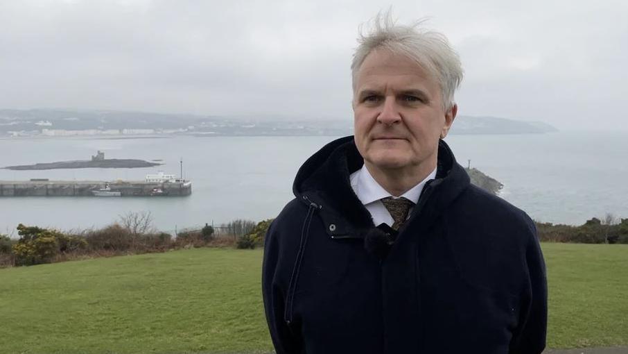 Diccen Sargent standing on the green hillside on Douglas Head with Douglas Bay in the background. He has grey hair and is wearing a white shirt and green tie, with a black coat.