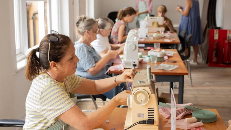 Women sit around sewing machines on tables in a community hall setting as part of a sewing group. The woman at the front has dark hair in a ponytail and is wearing glasses on top of her head. She is threading the machine. There are some older women sat near her. There are some women near the back stood up, helping the women with their project.