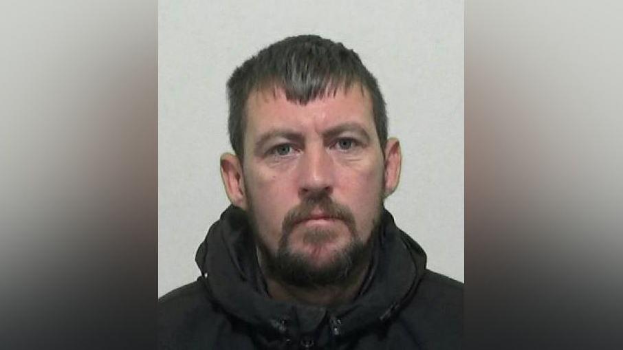 Police mugshot of Liam Doeg. He has black hair and a black beard and is wearing a black hooded coat.