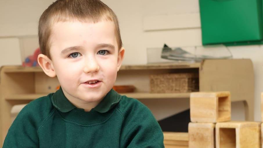 Lincoln Button is smiling at the camera, he has blue eyes, short brown hair and is wearing a dark green jumper over a dark green polo shirt. 