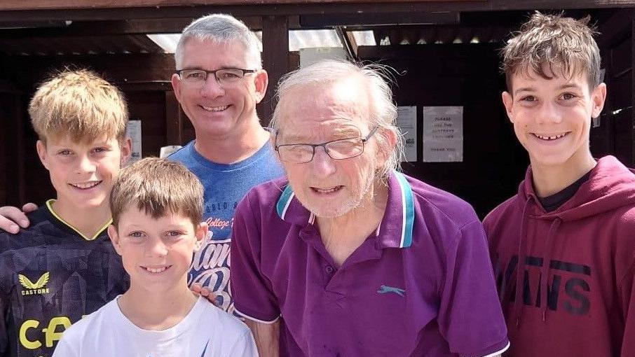 Two men and three boys wearing tshirts pose for a photograph in the sunshine