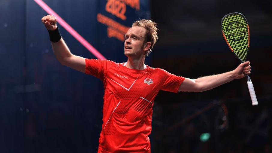 A squash player with blond hair in a red t-shirt holds his racquet up and punches the air.