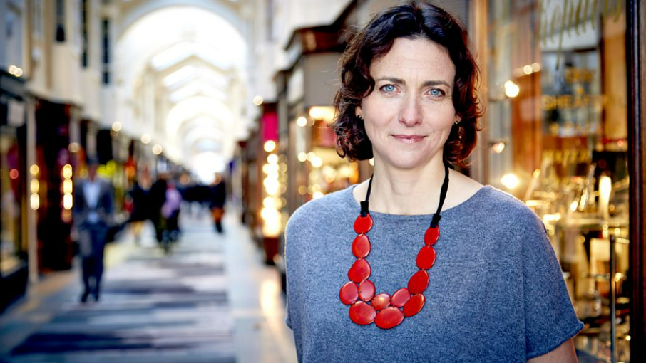 A head-and-shoulders shot of Dr Pamela Cox, wearing a red necklace and blue top, standing in a shopping arcade