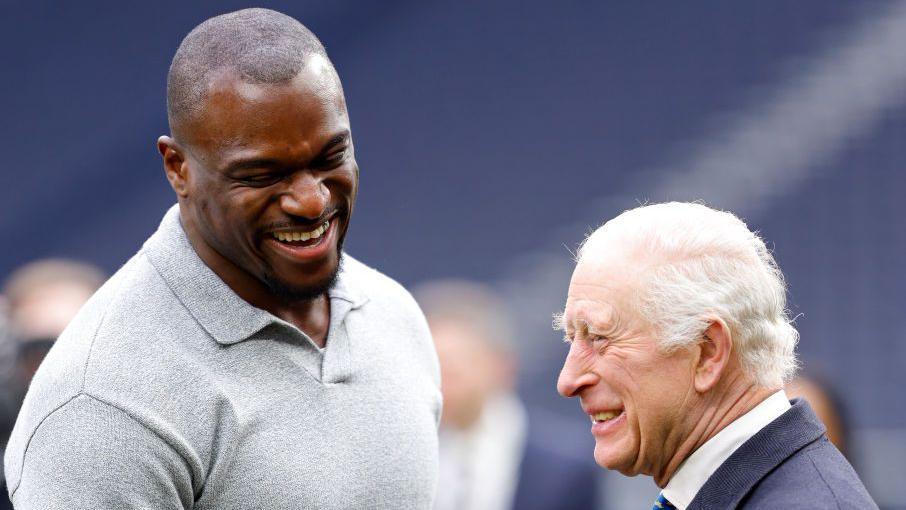 King Charles III and Efe Obada share a joke as they attend a demonstration by young people involved in The Huddle Project at Tottenham Hotspur Stadium in February 2025