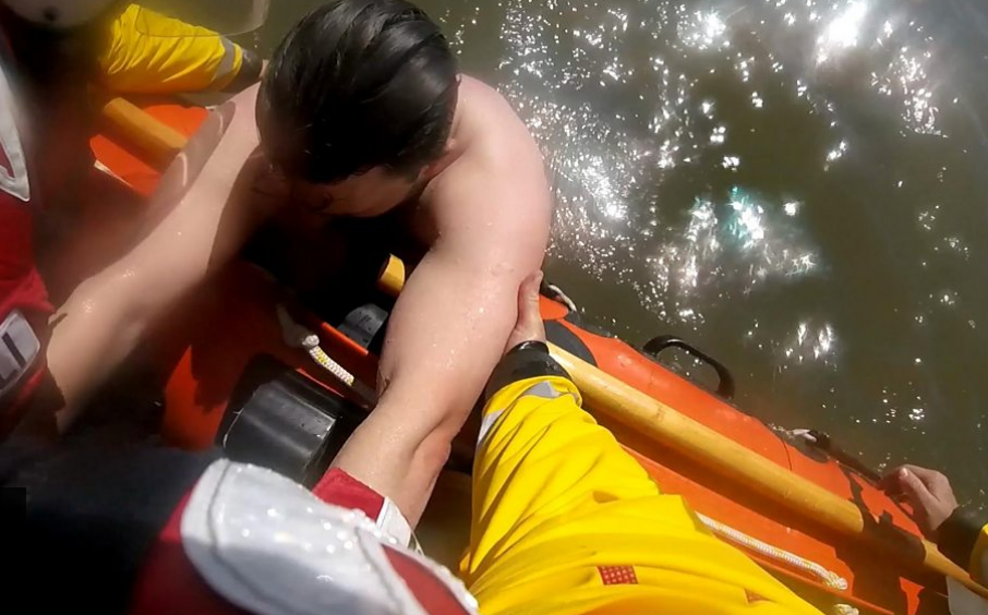 A man is pulled from the water onto a lifeboat. He is being lifted into the dinghy by his arms. An RNLI crewmember's leg, in yellow oilskins, is visible on the side of the orange RIB. 