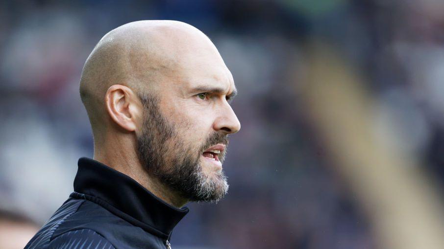 New Peterborough United manager Luke Williams looks on from the touchline during his time at Swansea City. He has a shaven head and a dark beard. He is wearing a navy blue tracksuit top.