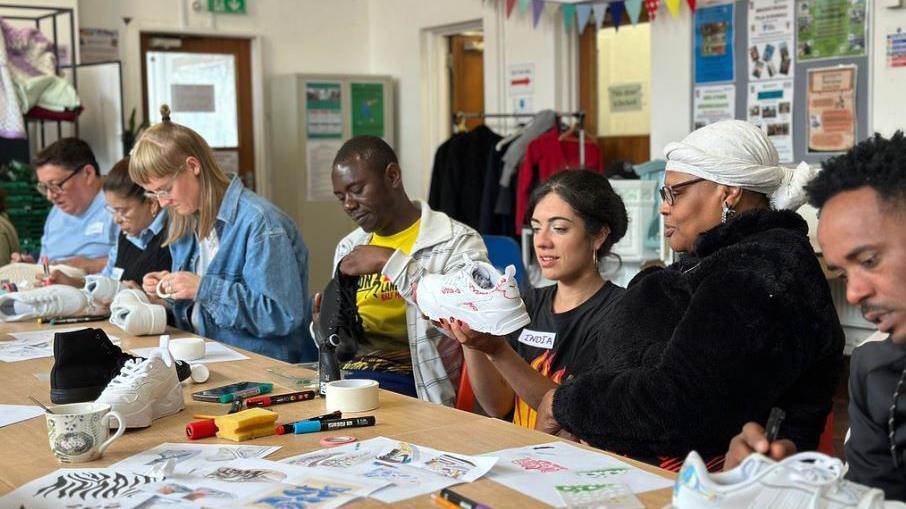 A group of people sit on one side of a table that is filled with paper, pens and art materials. All are intensely decorating a shoe each.