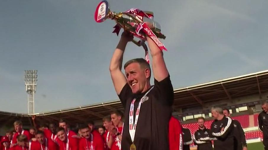 A man wearing a black polo shirt lifts a trophy above his head. He is wearing a medal around his neck. A group of men wearing red shirts stands behind him. They are on the pitch inside a stadium.