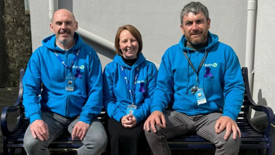 A woman sitting in between two men on a black bench. All three are wearing bright blue hoodies and smiling.