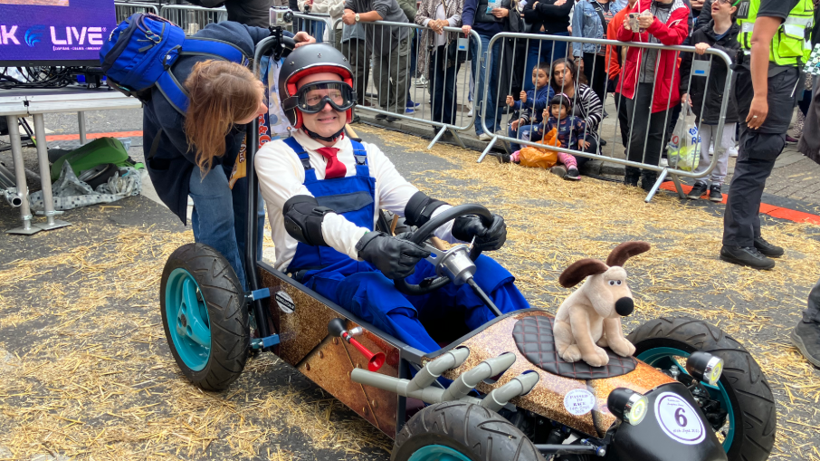 A person is dressed in a white shirt with a red tie and blue overalls. They have a black helmet and goggles on and are sat in a small kart which has a brown coloured teddy of Gromit from Wallace and Gromit on the front. 