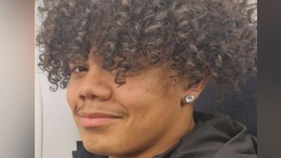 Isaac Brown in a photograph handed out by police. He has thick, curly hair and an ear stud, and he is wearing a black top. He is facing the side with his face turned to the camera, smilling.