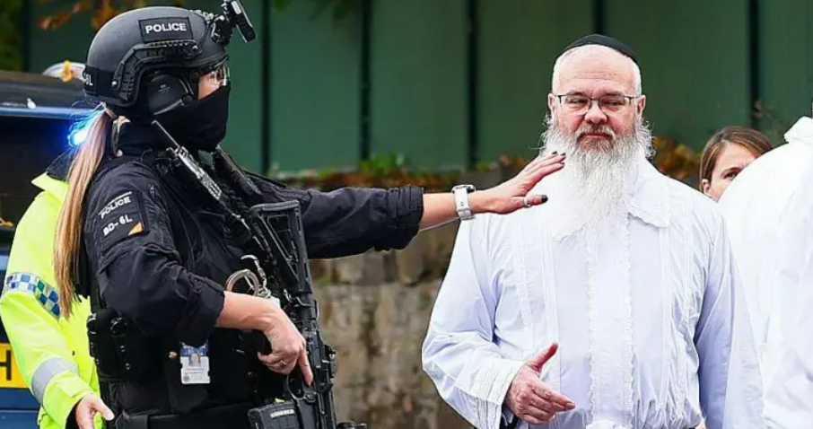 Rabbi Daniel Walker, wearing a white kittel, is standing next to an armed police officer. 