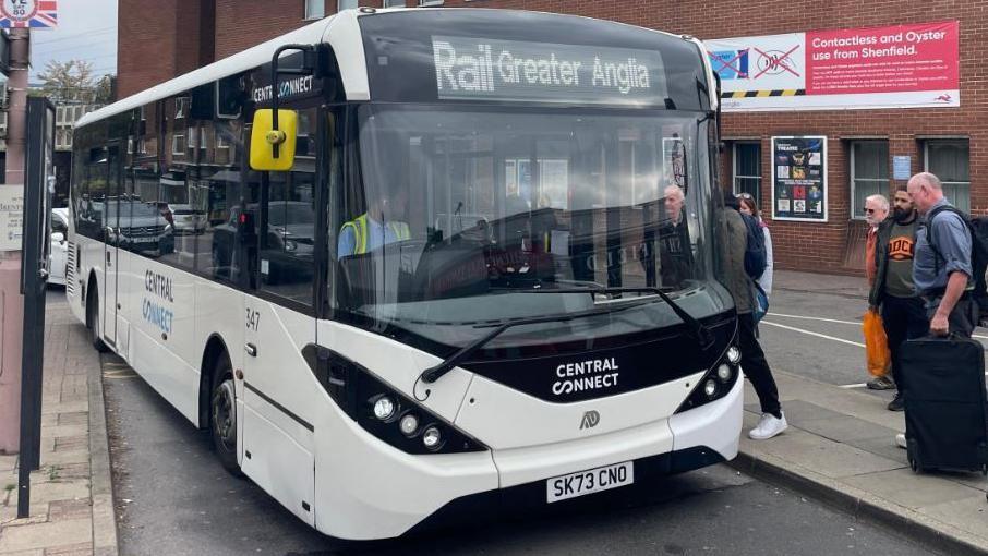 A white single-decker bus with the words 'Rail: Greater Anglia' on its sign. There is a queue of people waiting to get on.
