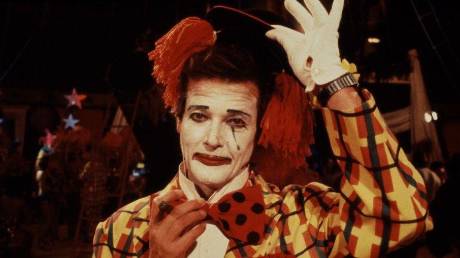 Roger Moore as James Bond dressed as a clown. He has a white painted face, red mouth, large red and black spotted bow tie and a yellow, black and orange checked suit. He is tipping his hat and holding a cigar.