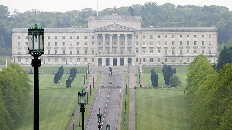 Stormont, a large stone building with numerous windows is positioned at the top of a hill. On either side of the road towards the building is grass and trees.